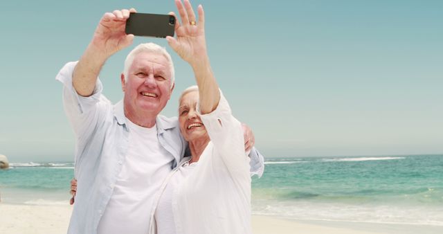 Senior couple enjoying a sunny day at the beach while taking a selfie with a smartphone. The ocean waves are visible in the background, creating a serene atmosphere. Perfect for concepts related to retirement, family vacations, happiness, and enjoying life.