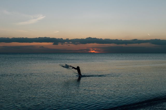 Silhouette of a person casting a fishing net into the calm sea at sunset. Sun is setting just above the horizon, creating a serene twilight atmosphere. Useful for concepts related to tranquility, peaceful fishing activities, evening leisure, and nature. Ideal for use in travel brochures, websites promoting outdoor activities, or posters highlighting serene coastal scenes.
