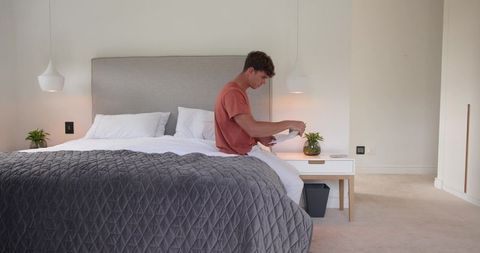 Young man sitting on bed folding shirt in bright minimalist bedroom with grey quilt
