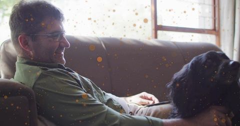 Middle-aged man relaxing on sofa petting large black dog by sunlit window, cozy home life