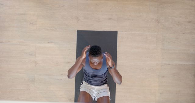 A young African American man is doing sit-ups on a yoga mat in a bright, minimalist room with wooden flooring. His workout attire suggests a commitment to fitness and a healthy lifestyle. Ideal for use in content promoting home fitness routines, gym workout programs, or health and wellness initiatives.