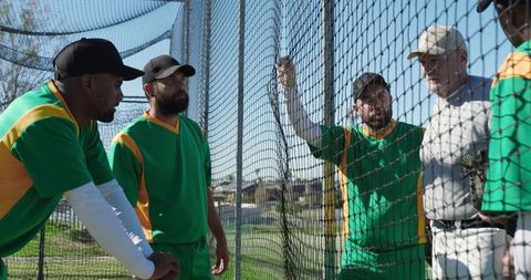 Diverse baseball team receiving coaching at batting cage fence during practice session