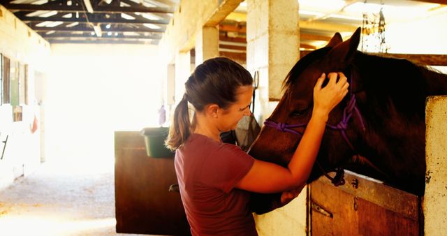 A young Caucasian woman gently interacts with a horse in a stable, with copy space. Her affectionate gesture towards the animal conveys a sense of care and connection between human and horse.
