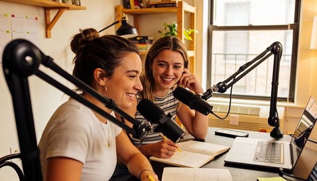 Two women recording podcast together at cozy home studio desk with microphones and laptop