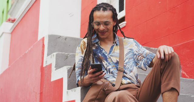 Smiling man with dreadlocks is sitting on stair, using smartphone in an urban area. He is dressed in casual clothing, wearing sunglasses, and looks relaxed and happy. Perfect for portraying urban lifestyle, modern fashion, technology use, social media engagement, or contemporary living.