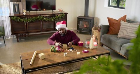African American man wrapping Christmas gifts at home wearing Santa hat by fireplace
