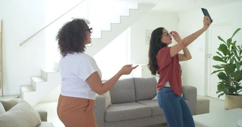 Diverse women posing and taking selfie with smartphone in modern bright living room