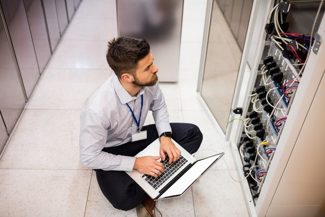 Technician kneeling on floor using laptop while analyzing server in data center. Ideal for illustrating IT support, network maintenance, and technology-related articles or advertisements. Can be used in blogs, websites, and marketing materials focused on information technology, technical support services, and data management.