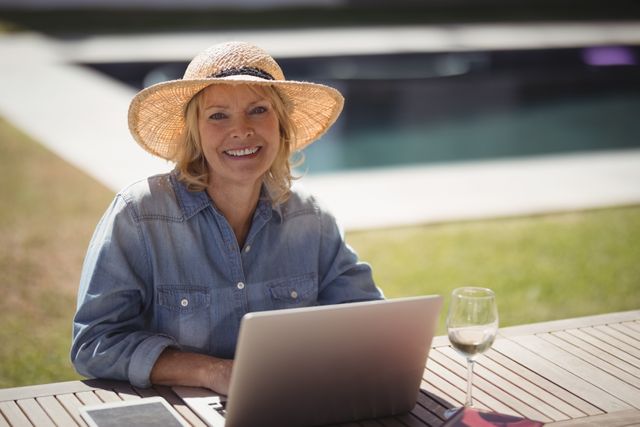 Senior woman sitting at a table in her garden, using a laptop and smiling. She is wearing a straw hat and casual clothing, with a glass of wine beside her. Ideal for use in articles about senior lifestyle, remote work, technology use among the elderly, and outdoor leisure activities.