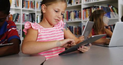 Elementary students using tablets in library girl in pink striped top learning technology