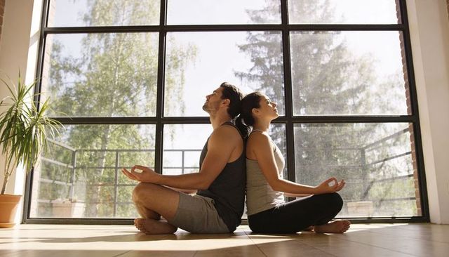 Back-to-back couple meditating in sunlit room with grid window for mindfulness