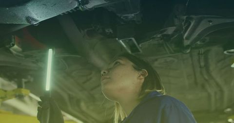 Female Mechanic Inspecting Car Undercarriage with Light in Workshop