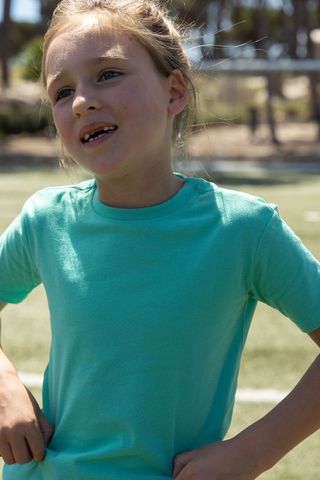 Young girl tugging turquoise t-shirt on soccer turf, standing and speaking candid portrait