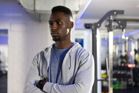 Focused man standing with crossed arms in modern gym wearing gray hoodie and smartwatch