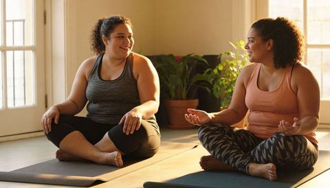 Two friends practicing mindfulness and chatting on yoga mats in sunlit home studio