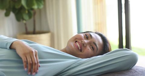 Mid adult asian woman reclining on sofa smiling wearing light blue top in sunlit room