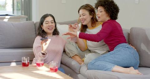 Diverse friends laughing while reaching for remote on cozy sofa with red drinks