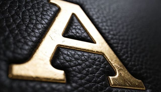 Gold letter a emblem on black pebbled leather macro showing patina, scratches and texture