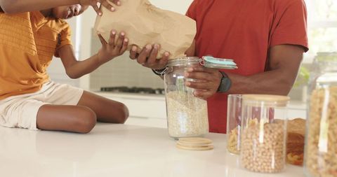 Father and son pouring dry oats from paper bag into glass jar on bright kitchen counter