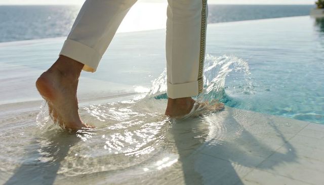Bare feet splashing at infinity pool edge in white cuffed trousers with sunlit ocean horizon