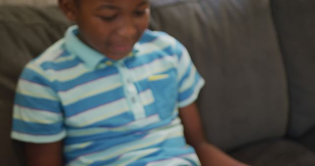 Young African American boy is reading digital tablet while sitting on couch. He is smiling, suggesting enjoyment. The boy wears a casual striped shirt. Useful for educational, technology, or family-oriented themes highlighting children's interactions with technology in home environments.