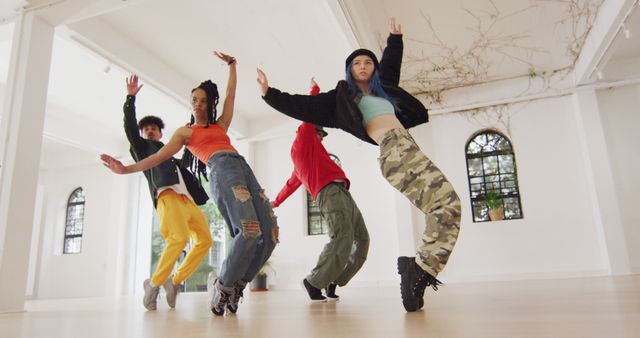 Diverse group of young adults performing a choreographed dance routine in a spacious, urban studio. They wear colorful, athletic attire as they express themselves through dynamic dance movements. Perfect for illustrating themes of teamwork, dance classes, contemporary dance, artistic expression, urban culture, and youth engagement.
