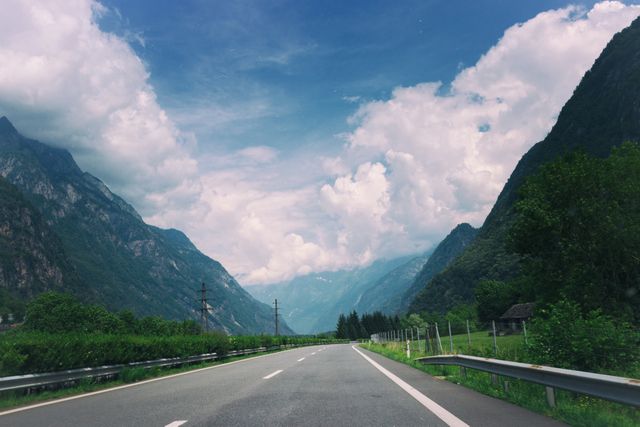 This stock photo depicts a scenic mountain highway surrounded by lush greenery and dramatic clouds. Ideal for travel blogs, adventure magazines, nature websites, and promotional material for road trips. Perfect background for outdoor adventure themed content.