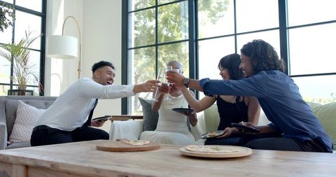 Group toasting with wine and pizza on sofa in bright modern living room celebrating together