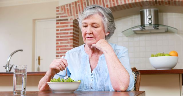 Senior woman wearing light blue blouse eating salad at kitchen table with glass of water and bowl of fruit. Brick and tile background emphasizes cozy home environment. Suitable for topics on healthy eating, aging well, homemaking, and nutrition advice for seniors.
