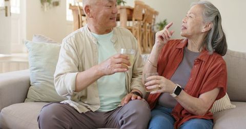Asian senior couple sitting on sofa sharing drink and enjoying warm conversation