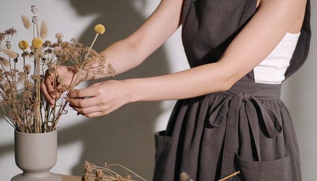 Woman arranging dried wildflower bouquet in ceramic pedestal vase, wearing grey apron
