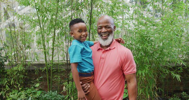 A loving grandfather holding his grandson while enjoying a day outdoor surrounded by lush greenery. Both are smiling joyfully, showcasing their close bond. Perfect for themes of family, multigenerational relationships, outdoor activities, and happiness. Can be used for family wellness, lifestyle, or summer activity promotions.