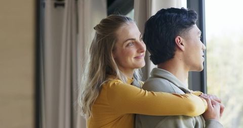 Couple embracing by window in modern apartment, woman hugging partner, cozy tender moment