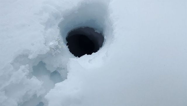 Circular hole cutting through packed snow on frozen lake revealing deep dark water void