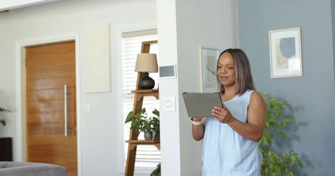 Woman holding tablet standing in sunlit modern living room with ladder shelf and plants