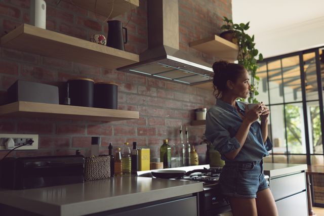 A woman is standing in a modern kitchen, holding a cup of coffee and appearing thoughtful. Shelves and kitchen appliances are visible in the background, giving the space a cozy and contemporary feel. Ideal for use in lifestyle blogs, home decor websites, and advertisements promoting modern kitchen designs or coffee-related products.