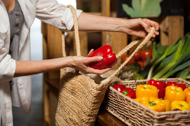 Woman selecting fresh bell peppers from a basket at an organic market. Ideal for use in content related to healthy eating, sustainable shopping, organic produce, and eco-friendly lifestyle. Perfect for blogs, articles, and advertisements promoting local markets and healthy living.
