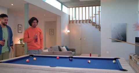 Friends playing pool in modern living room holding bottled drinks and cues