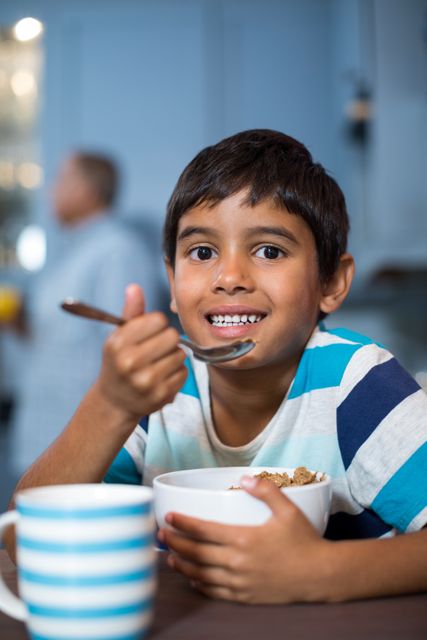 Boy enjoying a healthy breakfast of cereal at home, smiling and looking at the camera. Ideal for use in advertisements for breakfast foods, family lifestyle promotions, and healthy eating campaigns. Perfect for illustrating morning routines and family life in a kitchen setting.