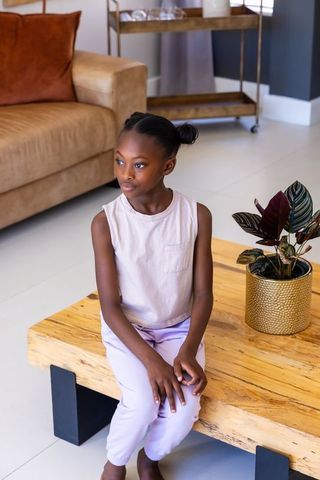 Girl sitting on light wood coffee table in minimalist sunlit living room, gazing pensively