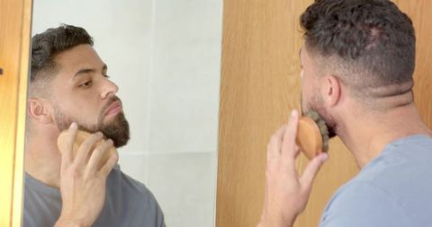 Man grooming beard in mirror with wooden brush morning self-care routine at home