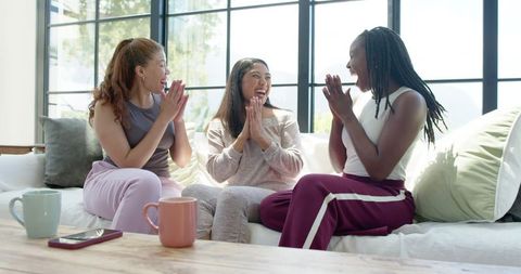 Three diverse women laughing, clapping and chatting in loungewear on bright modern sofa