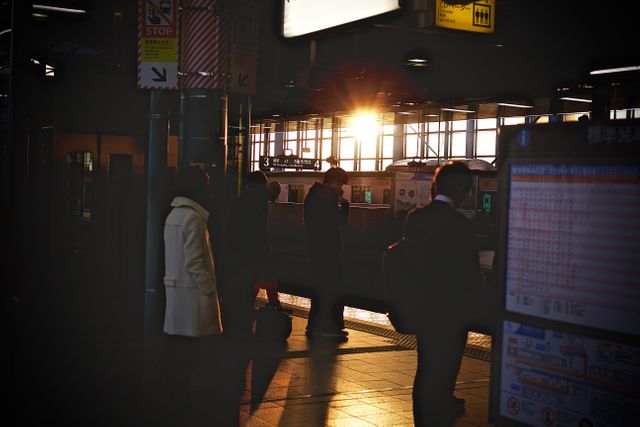 People are standing at a train station platform while the sun sets, casting a warm glow on the scene. This image can be useful for illustrating travel, commuting, urban life, transportation, and the hustle and bustle of city life campaigns or articles.