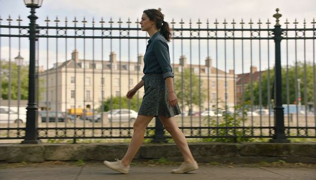 Woman walking along iron fence in denim shirt and patterned skirt, casual urban style
