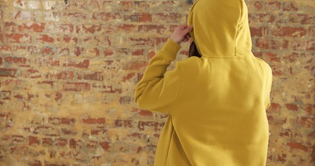 Person standing with back to camera wearing yellow hoodie, adjusting hood in a casual manner. This can be used for fashion blogs, urban lifestyle magazines, street style catalogs, or advertisements featuring modern casual wear or urban environment themes.