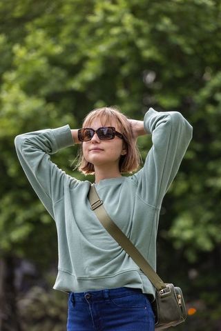 Young woman relaxing in park wearing sunglasses and sage sweatshirt with crossbody bag