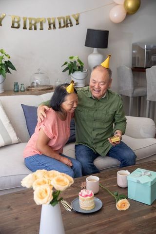 Asian senior couple celebrating birthday at home on sofa sharing cake, gift and smiles