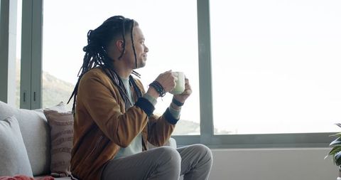 Man with long dreadlocks holding ceramic mug sitting on sofa by bright window morning calm