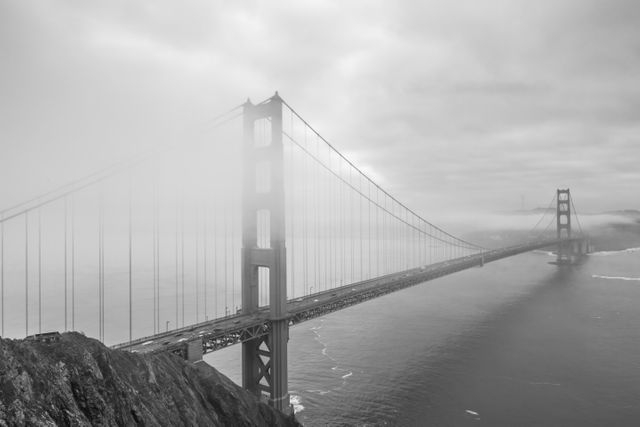 Depicting a fog-obscured Golden Gate Bridge under a cloudy sky, this dramatic black and white shot captures the majestic structure stretching across San Francisco Bay. Suitable for travel blogs, architectural features, weather-related content, and scenic photo collections.