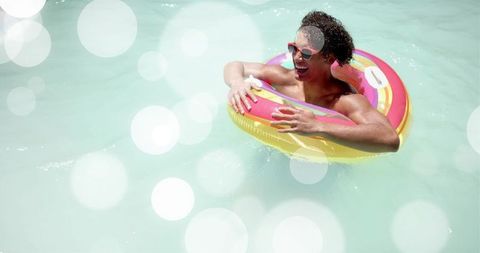 Man floating on doughnut inflatable in turquoise pool wearing red sunglasses, laughing